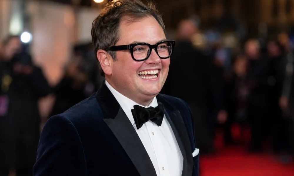 Alan Carr smiling in a formal tuxedo at a red-carpet event for an article about his 2026 net worth.