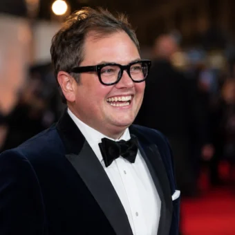 Alan Carr smiling in a formal tuxedo at a red-carpet event for an article about his 2026 net worth.