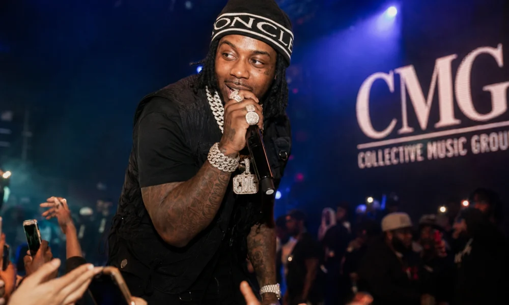 EST Gee performing live at a CMG Records concert with microphone, diamond jewelry, and crowd under blue stage lights.