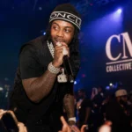 EST Gee performing live at a CMG Records concert with microphone, diamond jewelry, and crowd under blue stage lights.