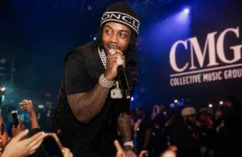 EST Gee performing live at a CMG Records concert with microphone, diamond jewelry, and crowd under blue stage lights.