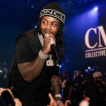 EST Gee performing live at a CMG Records concert with microphone, diamond jewelry, and crowd under blue stage lights.