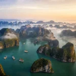 Aerial view of Ha Long Bay limestone karsts rising from emerald water at golden hour with mist and cruise boats in northern Vietnam