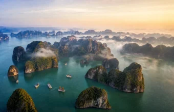 Aerial view of Ha Long Bay limestone karsts rising from emerald water at golden hour with mist and cruise boats in northern Vietnam