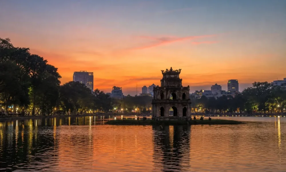 Turtle Tower silhouetted at dusk on Hoan Kiem Lake in Hanoi, Vietnam, with orange sunset reflections on the water.