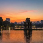 Turtle Tower silhouetted at dusk on Hoan Kiem Lake in Hanoi, Vietnam, with orange sunset reflections on the water.