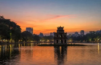 Turtle Tower silhouetted at dusk on Hoan Kiem Lake in Hanoi, Vietnam, with orange sunset reflections on the water.