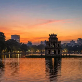Turtle Tower silhouetted at dusk on Hoan Kiem Lake in Hanoi, Vietnam, with orange sunset reflections on the water.