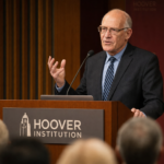 Speaker at a Hoover Institution-style podium during a public forum, used to illustrate Howard Lorber net worth estimates.