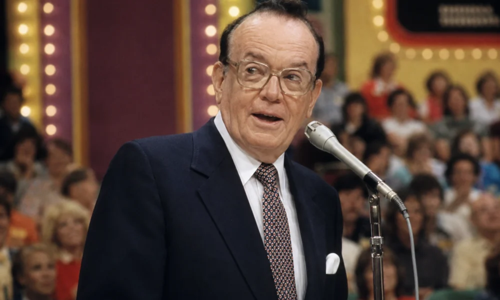 Johnny Olson speaking at a Price Is Right microphone in a vintage TV studio audience setting