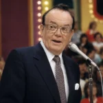 Johnny Olson speaking at a Price Is Right microphone in a vintage TV studio audience setting