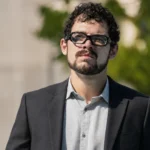 Rodrigo Blankenship wearing glasses and a dark suit outdoors.