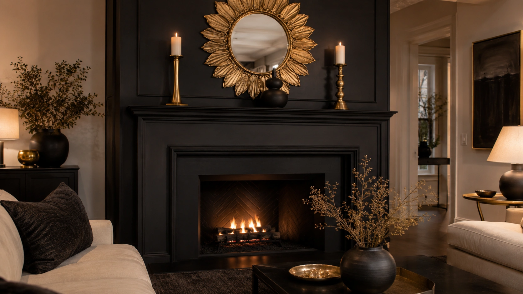 All-black fireplace surround with gold sunburst mirror, brass candleholders and cream seating in a moody living room.