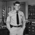 Andy Griffith in a black-and-white sheriff office scene with desk, flag, and rifle rack