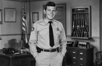 Andy Griffith in a black-and-white sheriff office scene with desk, flag, and rifle rack
