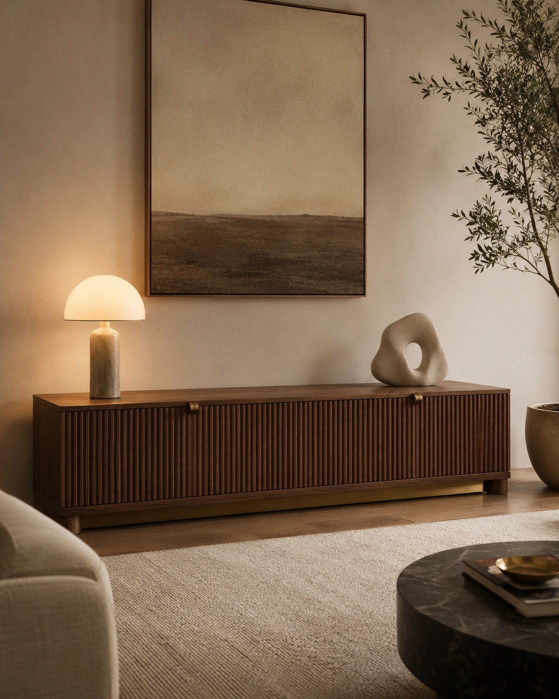 Fluted wood storage console with brass hardware, clean lines, warm lamp, wall art, and indoor olive tree.