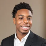 Isaiah Dickerson smiling in a black suit and white shirt against a neutral indoor background.
