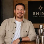 Jordan Childs smiling beside Shine Craft Vessel Co. stainless steel growlers with a branded sign in the background