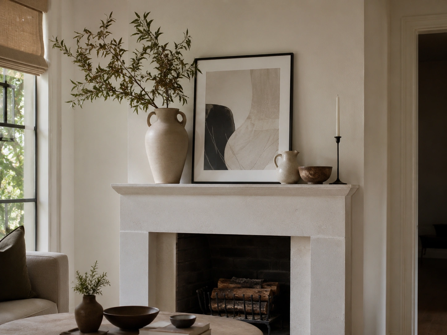 Layered mantel artwork behind sculptural vases, a wooden bowl, and a black taper candle in a neutral living room.