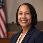 Neysa Fligor smiling in a navy blazer inside a public meeting chamber with a U.S. flag behind her