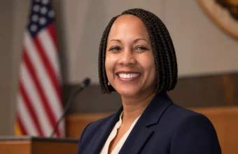 Neysa Fligor smiling in a navy blazer inside a public meeting chamber with a U.S. flag behind her