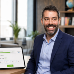 Paul Mishkin seated in an office beside a tablet showing an IXL math question