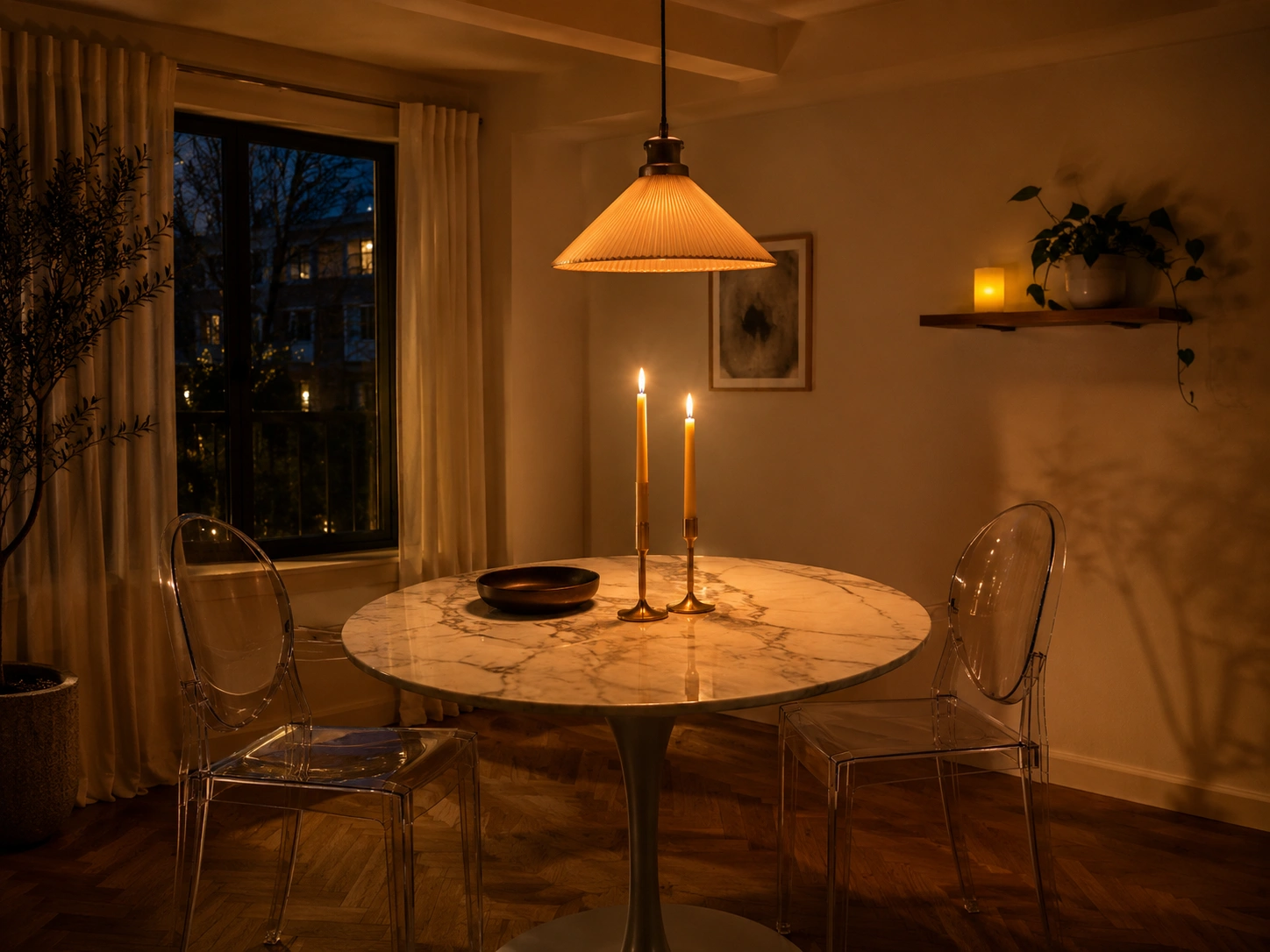 Small candlelit dining room with warm pendant light, round marble table, and clear ghost chairs.