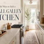 Small galley kitchen with cream cabinets, brass hardware, runner rug, and sunlight from a back window.