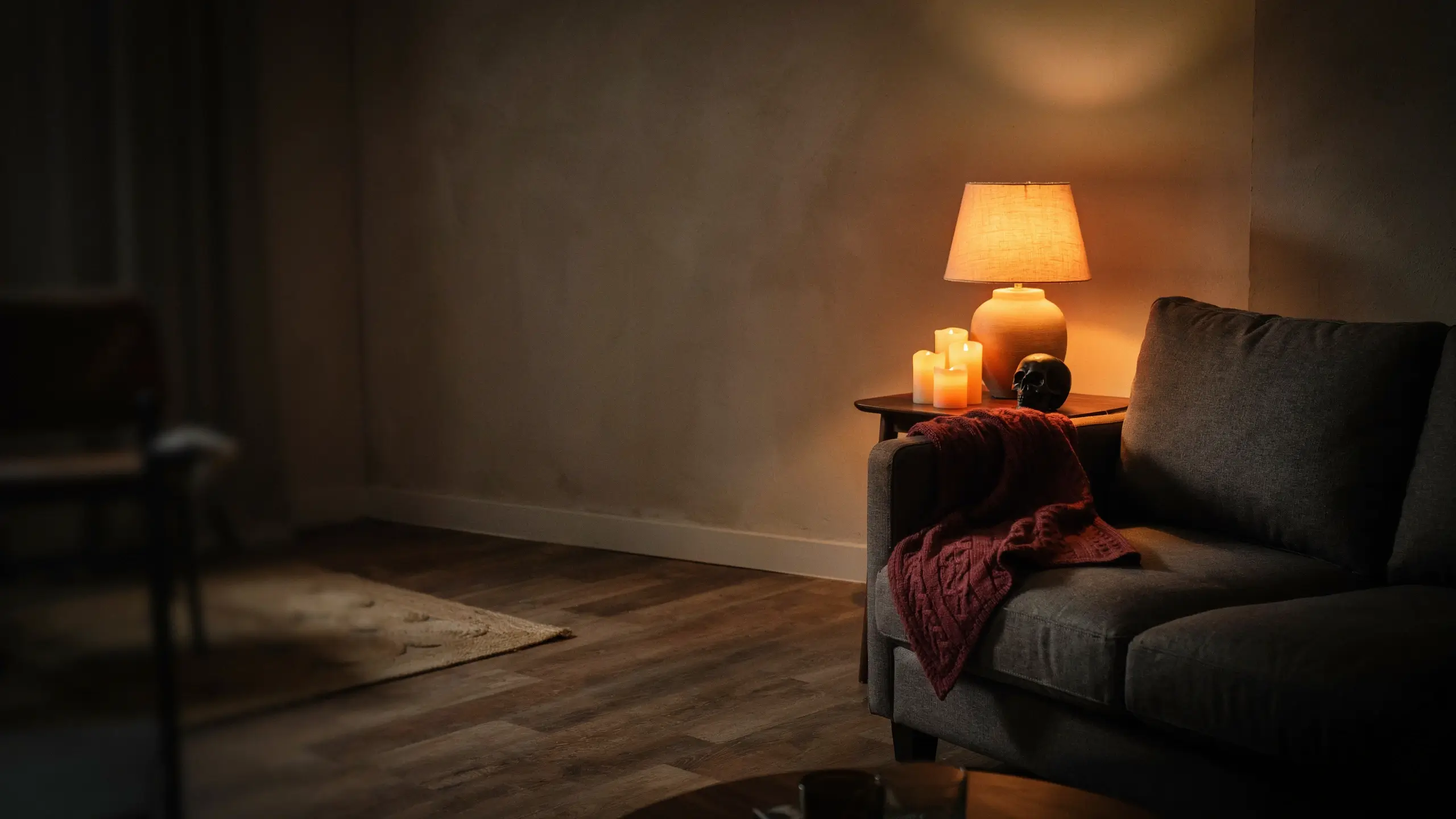 Cozy living room corner showcasing a warm orange ambient light setup inspired by a light string swap, featuring a glowing ceramic table lamp, three lit candles, and a black skull decor piece resting on a side table next to a grey sofa with a maroon knit throw blanket. 