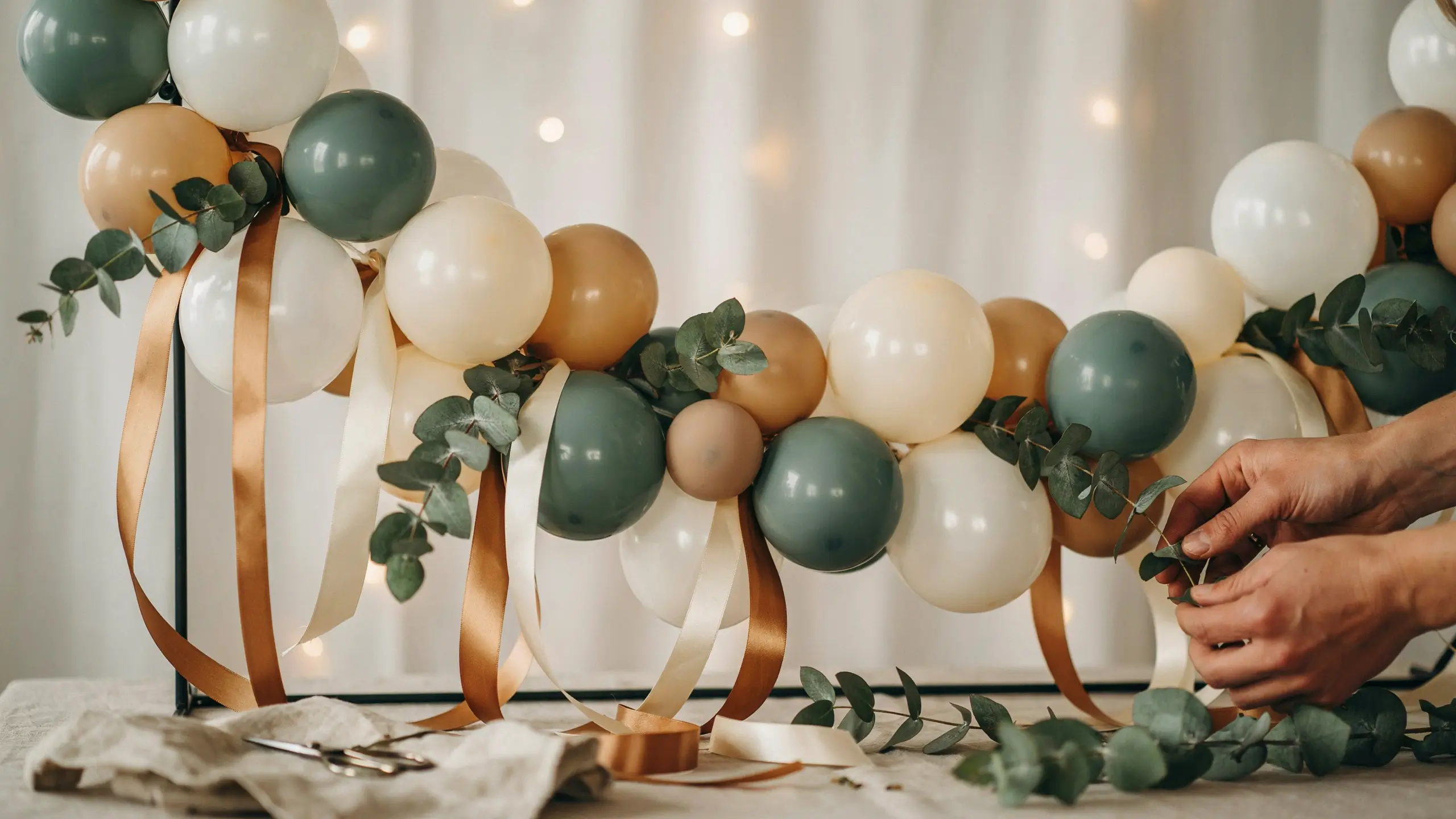 Hands weaving fresh eucalyptus sprigs and wide satin ribbons into a sage green, cream, and tan balloon garland as the final step of boho event decoration