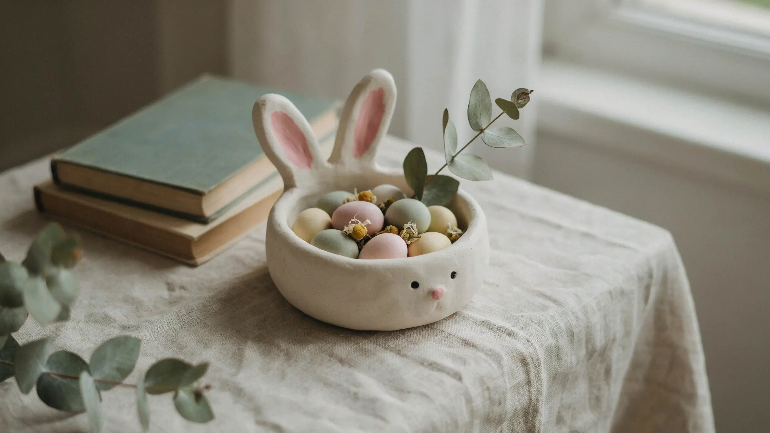 Handmade air-dry clay bunny bowl with pink ears, filled with pastel Easter eggs, dried flowers, and fresh eucalyptus leaves, sitting on a linen tablecloth beside stacked books. 
