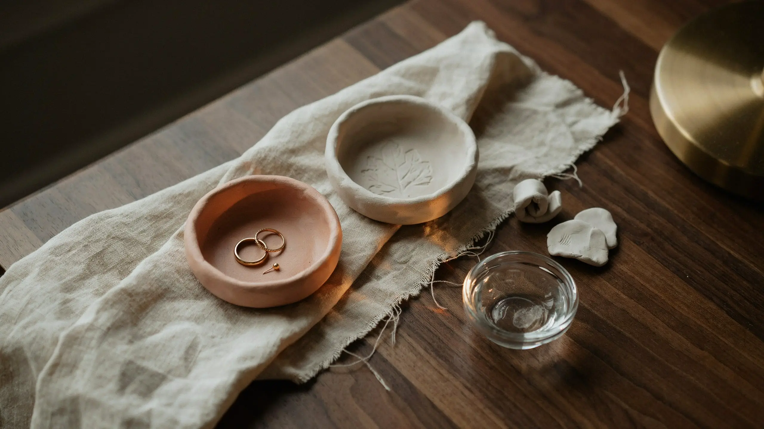 Two handmade air-dry clay trinket dishes on a linen cloth, one peach-toned dish holding a pair of gold wedding rings and a second off-white dish with a stamped leaf design.