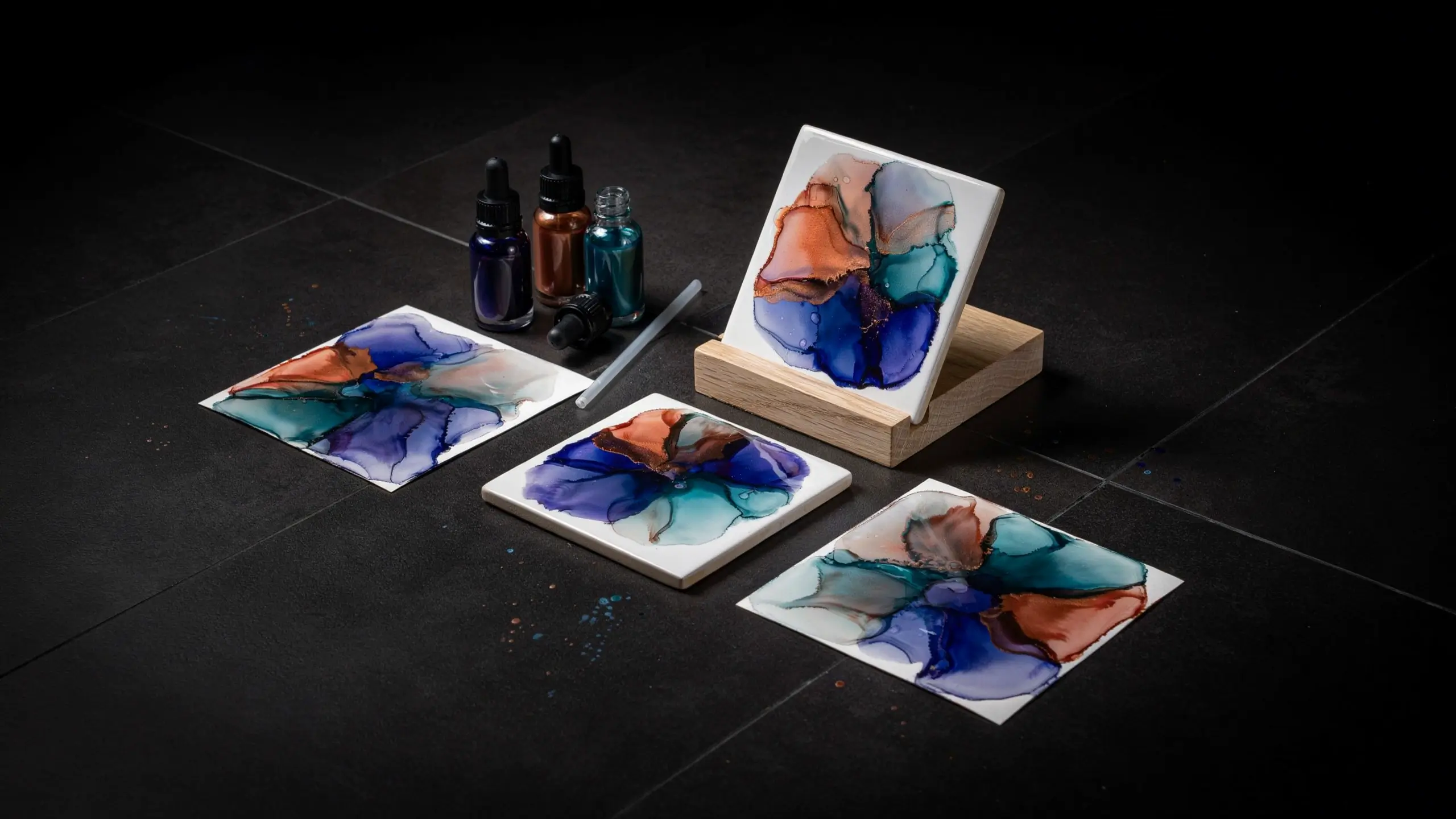 Square ceramic tiles and art panels displaying abstract alcohol ink pour art in deep blue, teal, and metallic copper tones, arranged flat and upright on a wooden display stand alongside small ink bottles and a stirrer on a dark slate work surface. 