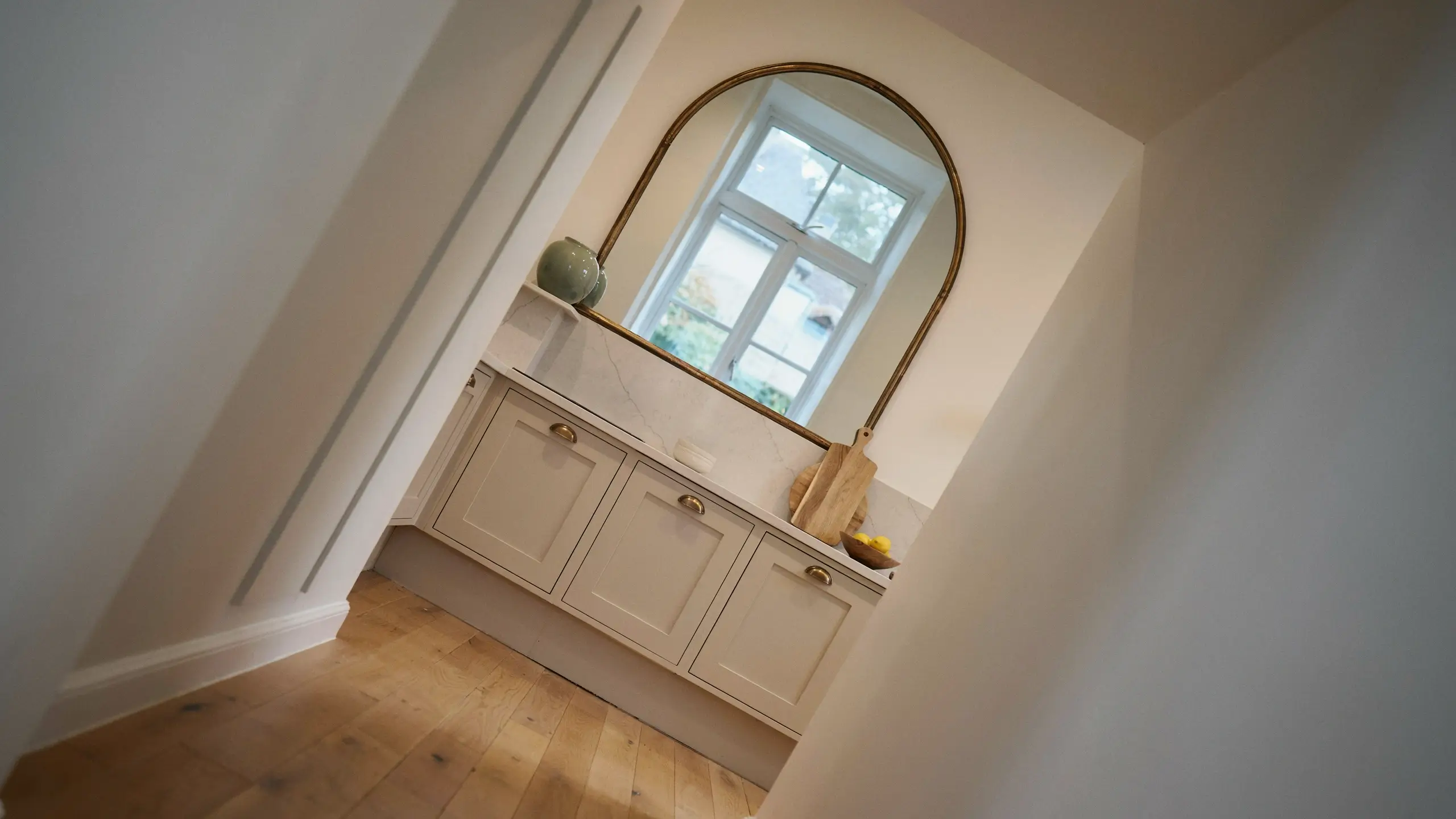 Arched brass mirror above cream kitchen cabinets reflecting natural window light