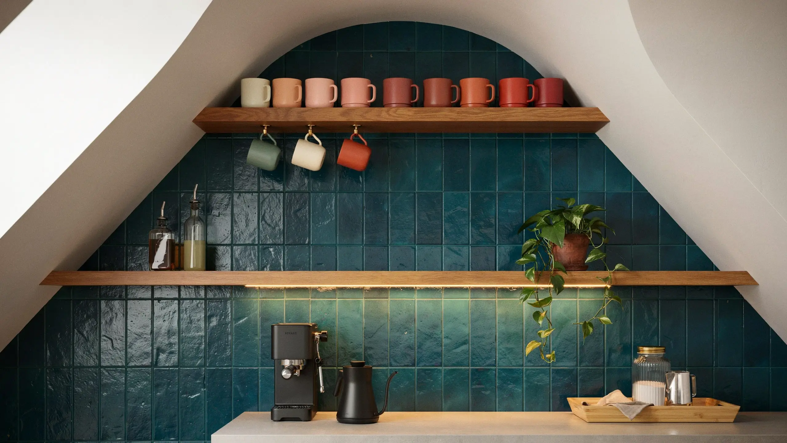 Stylish arched alcove coffee station with teal tiles, two wooden shelves displaying assorted mugs, a black espresso machine, a gooseneck kettle, and a hanging pothos plant.