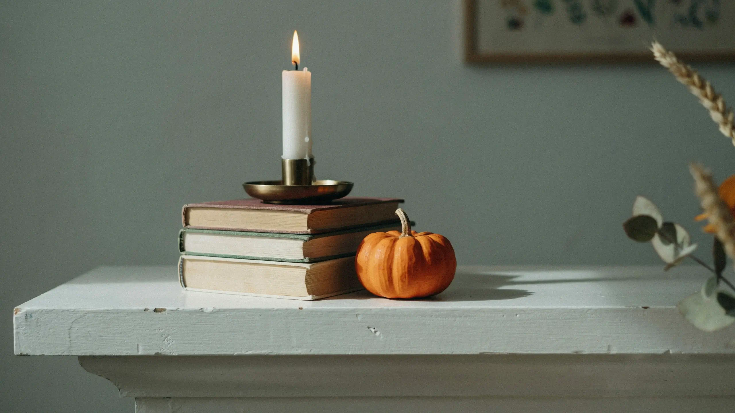 A white taper candle glowing on a brass holder, placed on a stack of three vintage books used as instant risers, next to a small orange pumpkin on a white wooden mantelpiece for cozy fall decor.