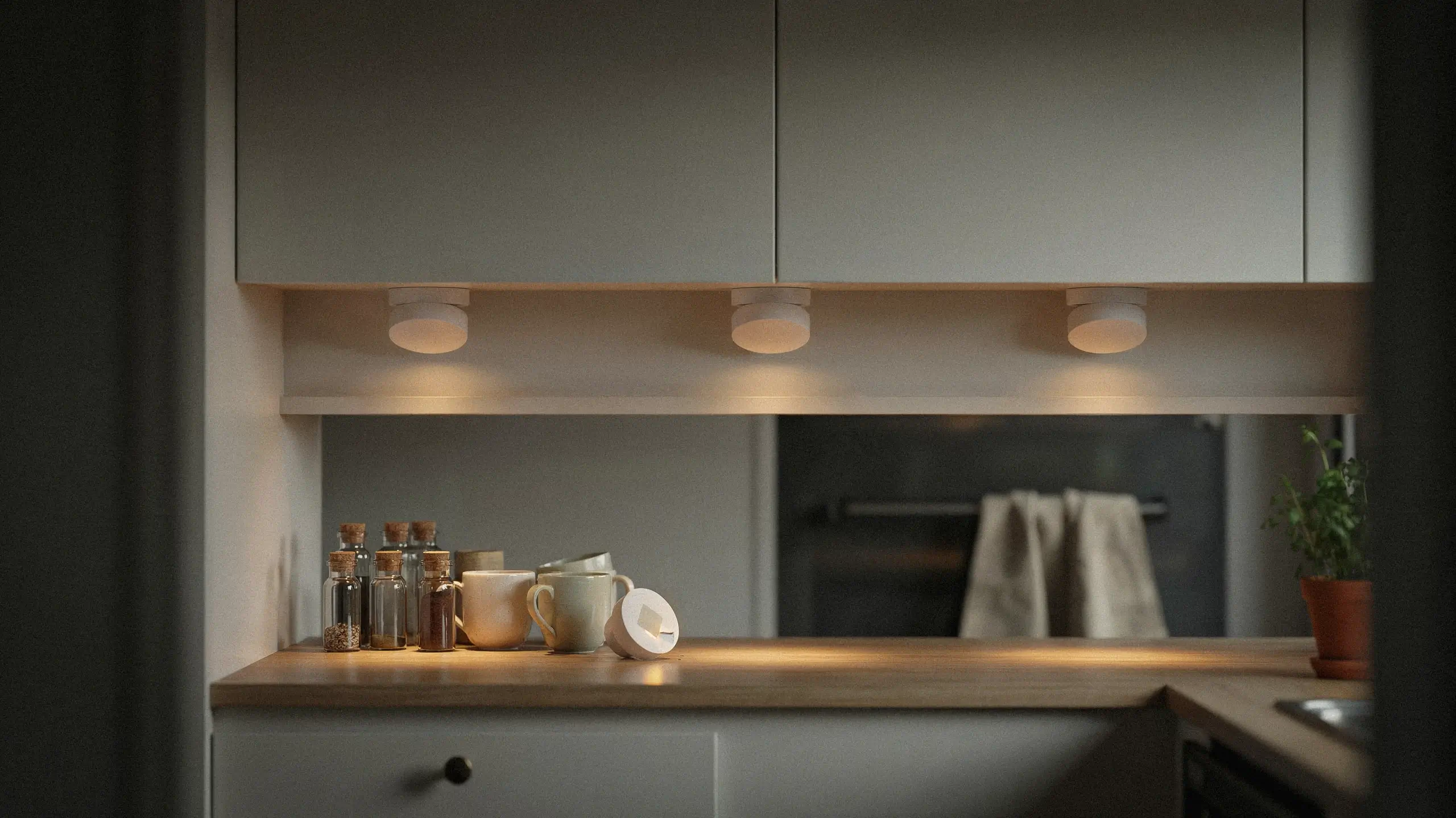 Wireless battery-powered puck lights mounted underneath white kitchen cabinets illuminating a wooden countertop with glass spice jars, ceramic mugs, and a potted plant, demonstrating easy under-shelf lighting solutions