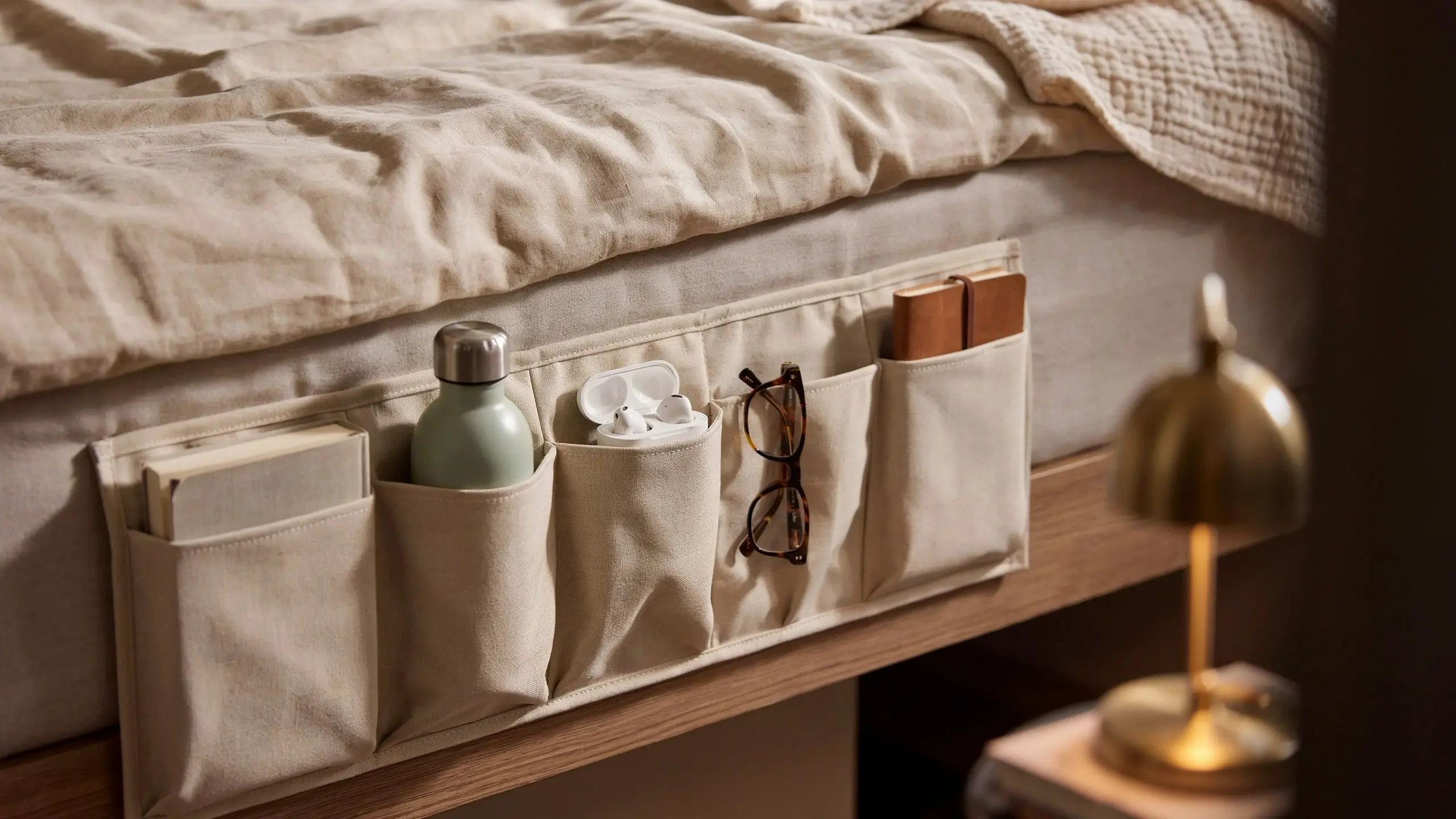 Beige fabric bedside caddy organizer with multiple pockets storing a book, water bottle, earbuds, glasses, and journal, attached to a wooden bed frame.