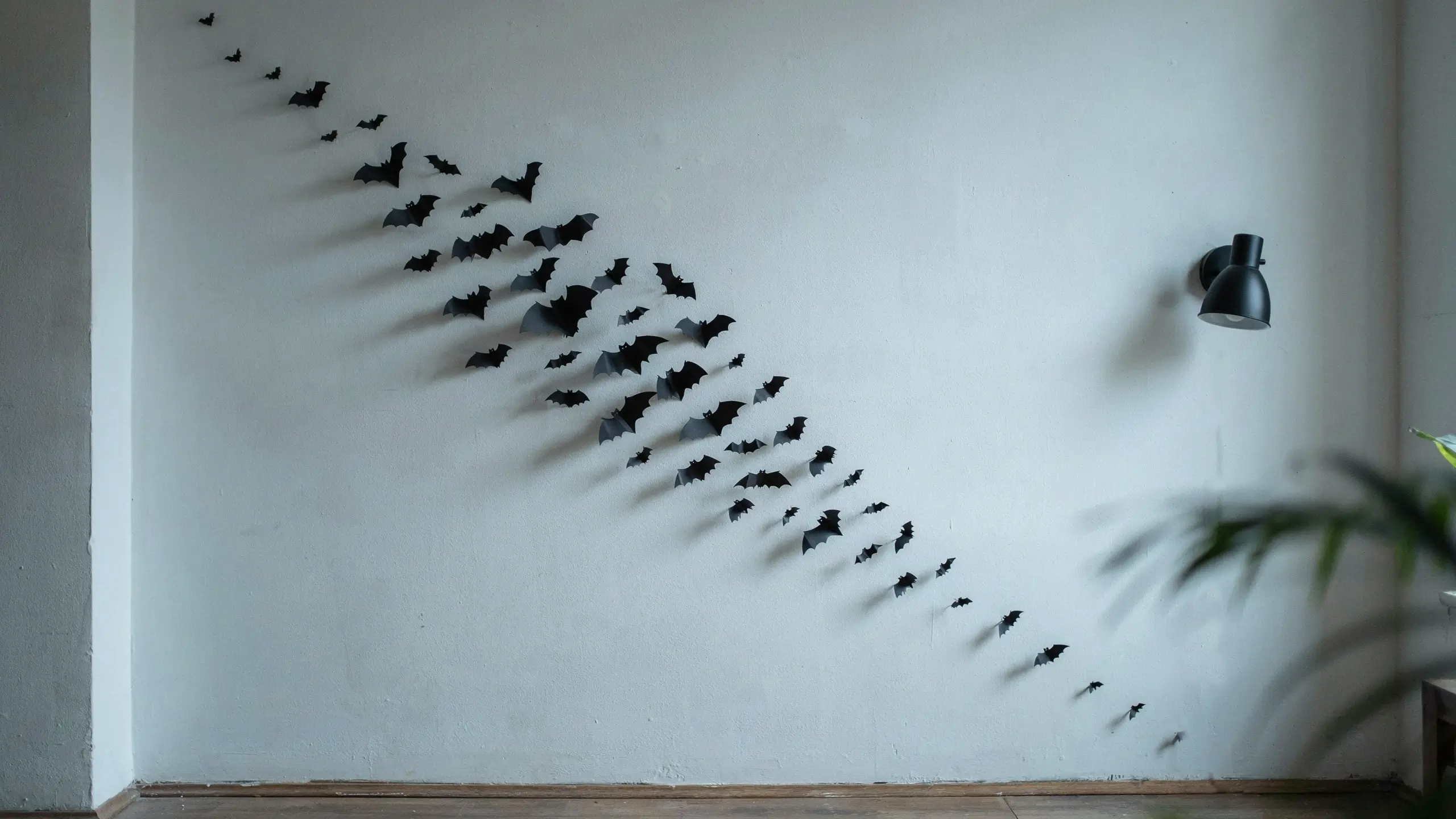 Minimalist black bat wall swarm decoration arranged in a sweeping flight pattern on a white wall, complemented by a black wall lamp and greenery for spooky home decor. 