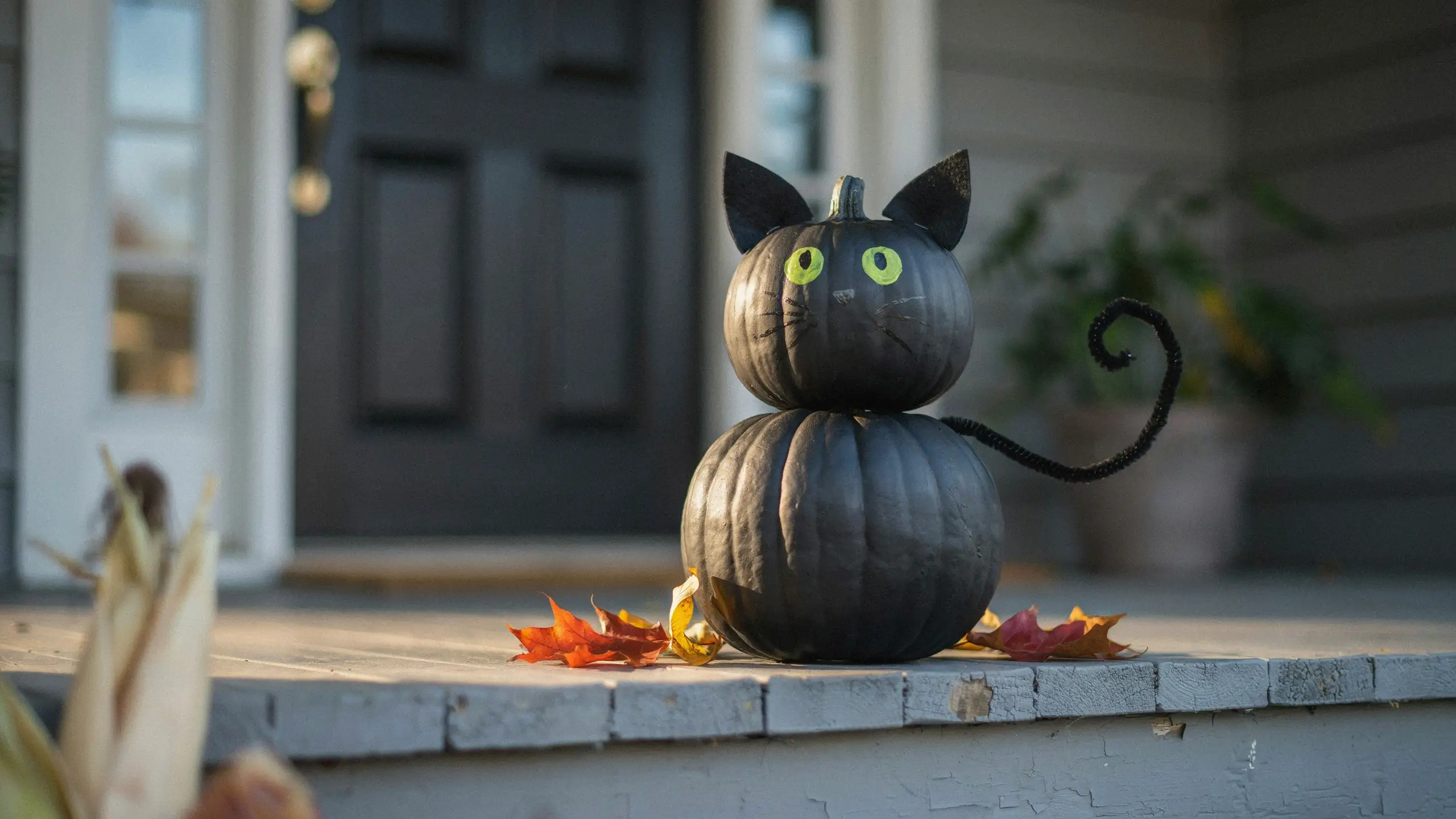 Stacked black pumpkins painted as a Halloween cat with yellow eyes and paper ears, resting on a wooden porch with scattered autumn leaves. 