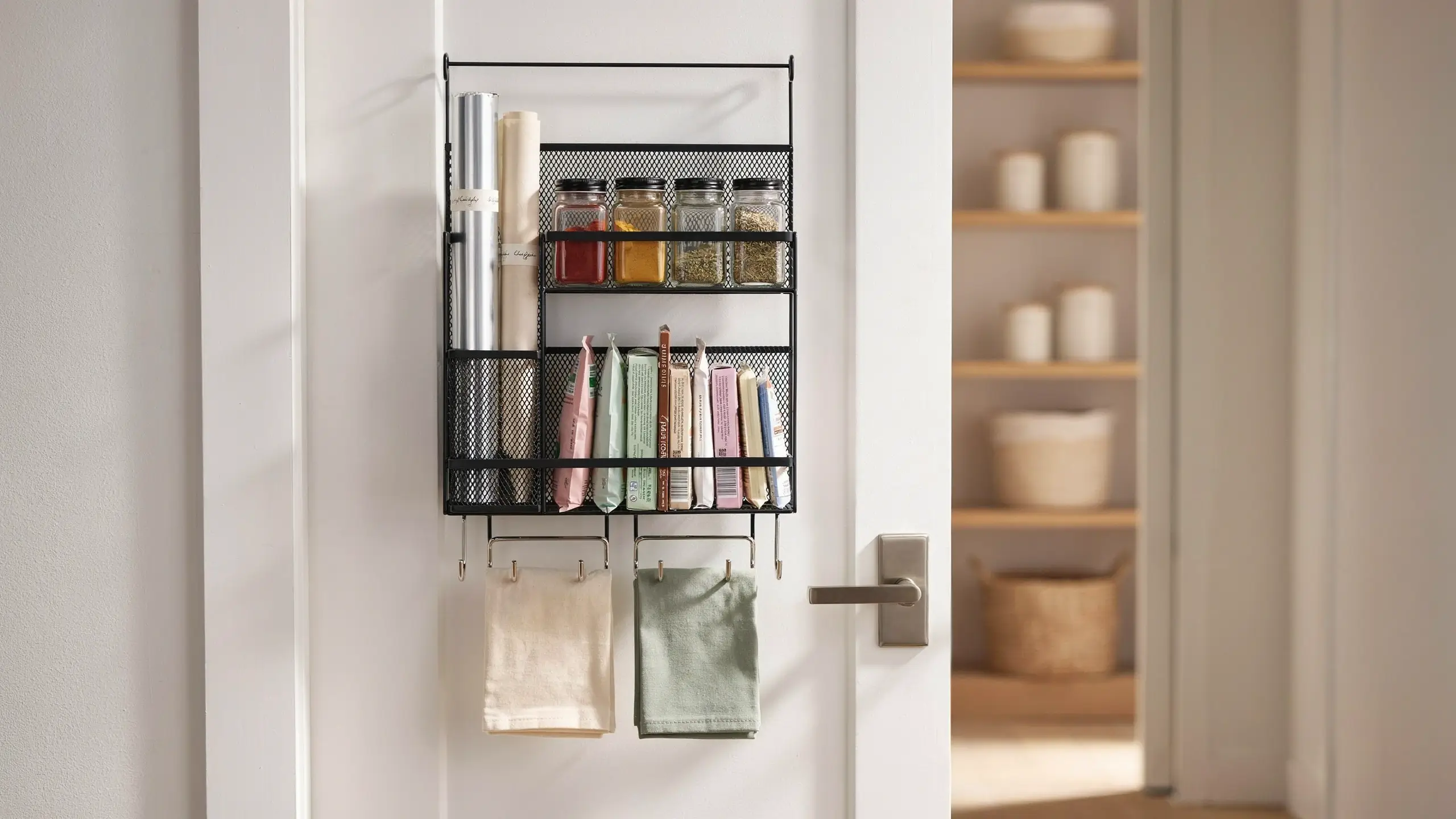 Black wire mesh over-the-door storage rack holding glass spice jars, kitchen rolls, and hanging cream and sage green dish towels on a white pantry door. 