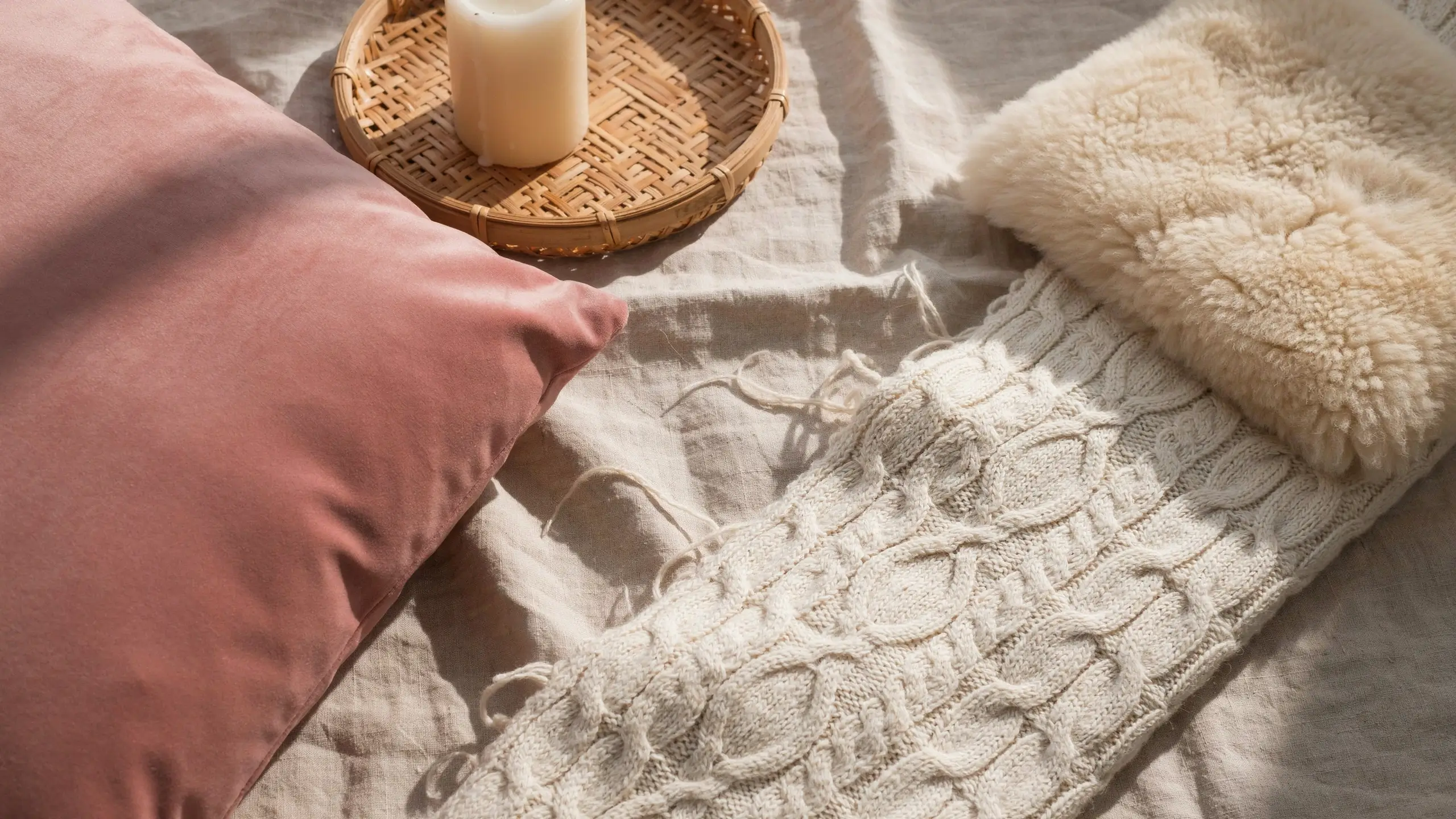 Cozy boho bedroom styling with a dusty rose pink throw pillow, a textured cream cable-knit blanket, a fluffy white faux fur accent, and a woven bamboo tray holding a white candle, all resting on rumpled linen sheets bathed in warm, natural sunlight.