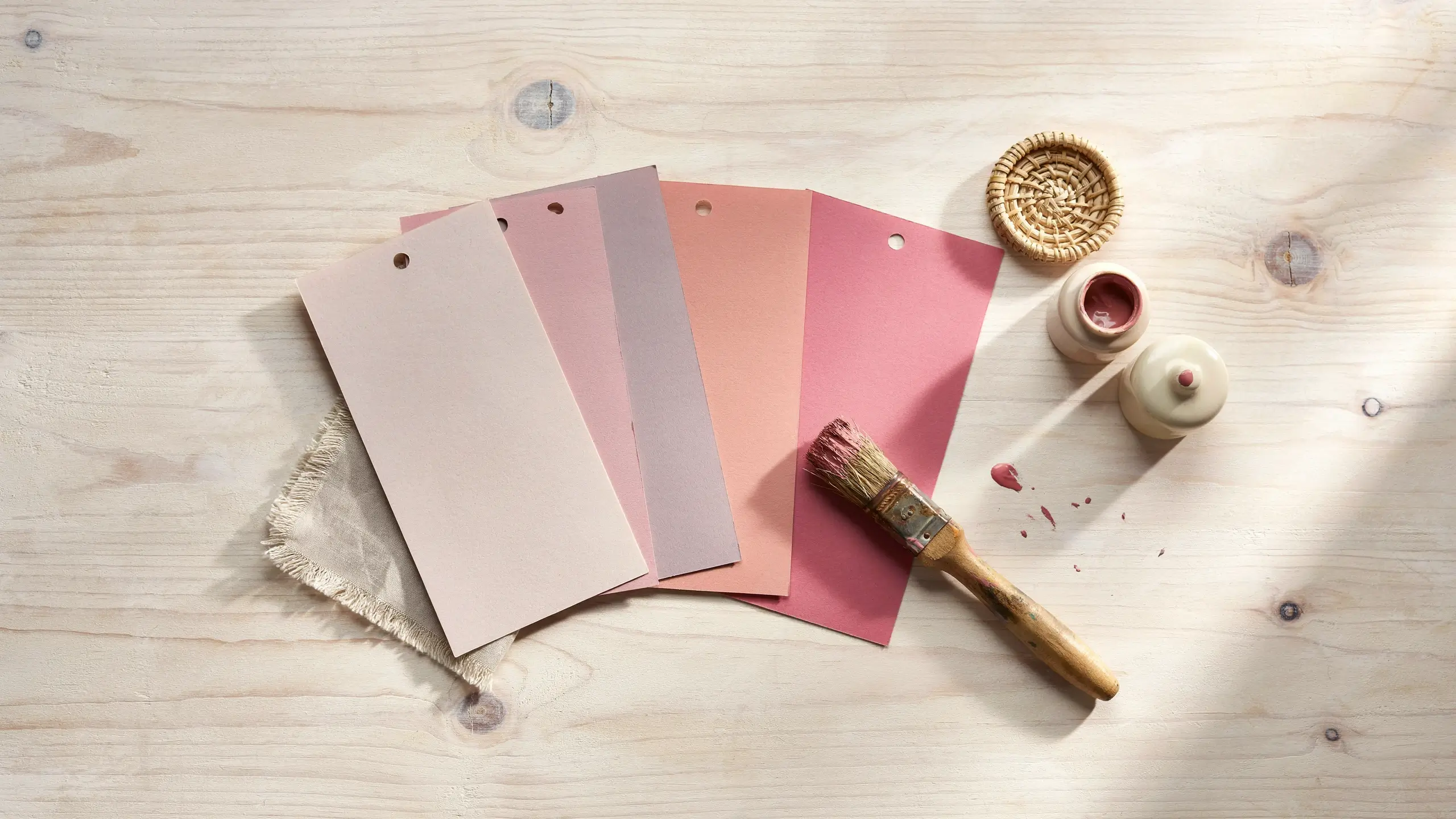 A flat lay of dusty pink, blush, and rose paint swatch cards on a light wooden table, arranged next to a paintbrush and an open jar of paint, ideal for selecting an interior wall color. 