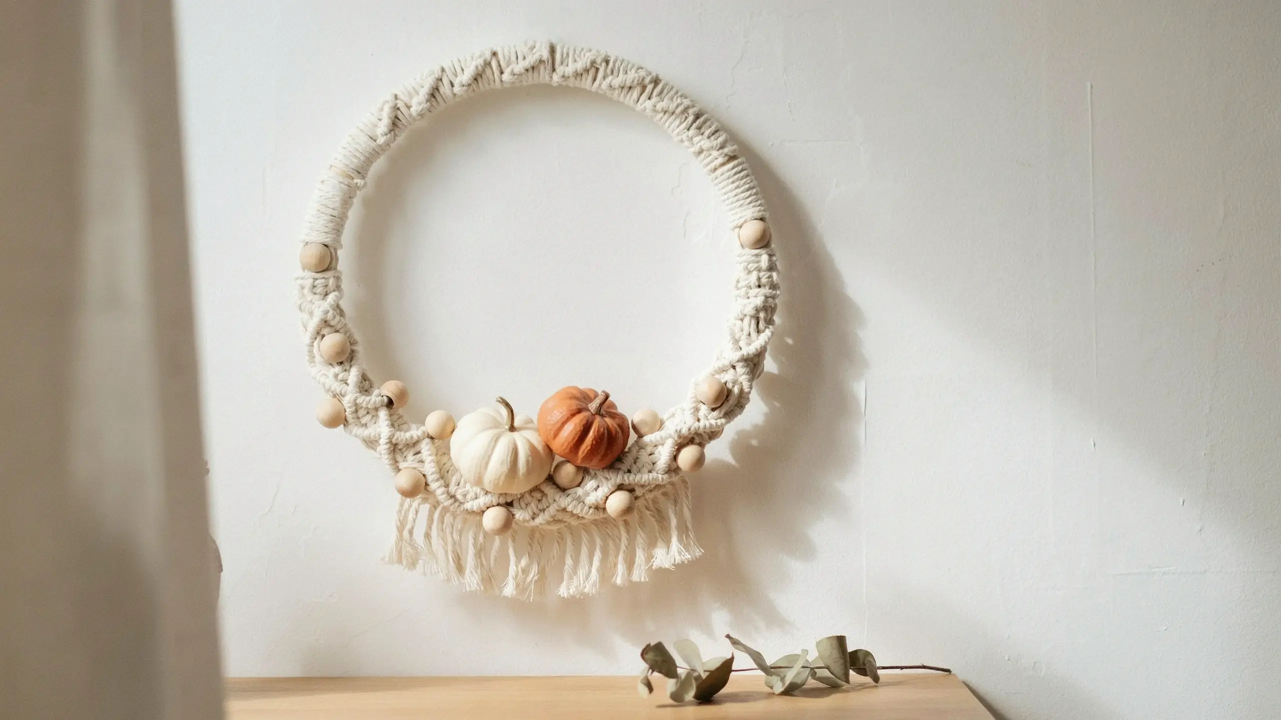 "Minimalist boho macrame wreath with wooden beads and mini pumpkins hanging on a white wall."