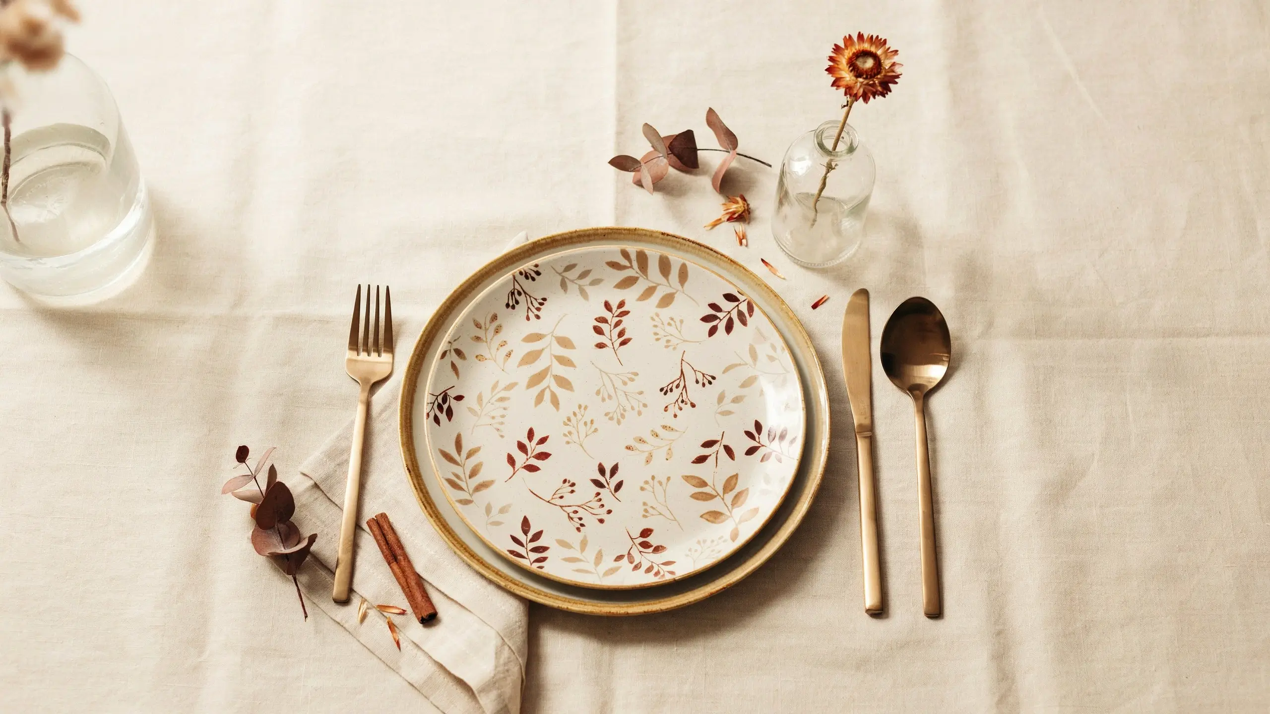 Two stacked white ceramic plates featuring rust and beige botanical leaf patterns, flanked by matte gold cutlery, a fabric napkin with dried eucalyptus, and a small glass vase holding a dried orange strawflower, arranged on a natural beige linen tablecloth.