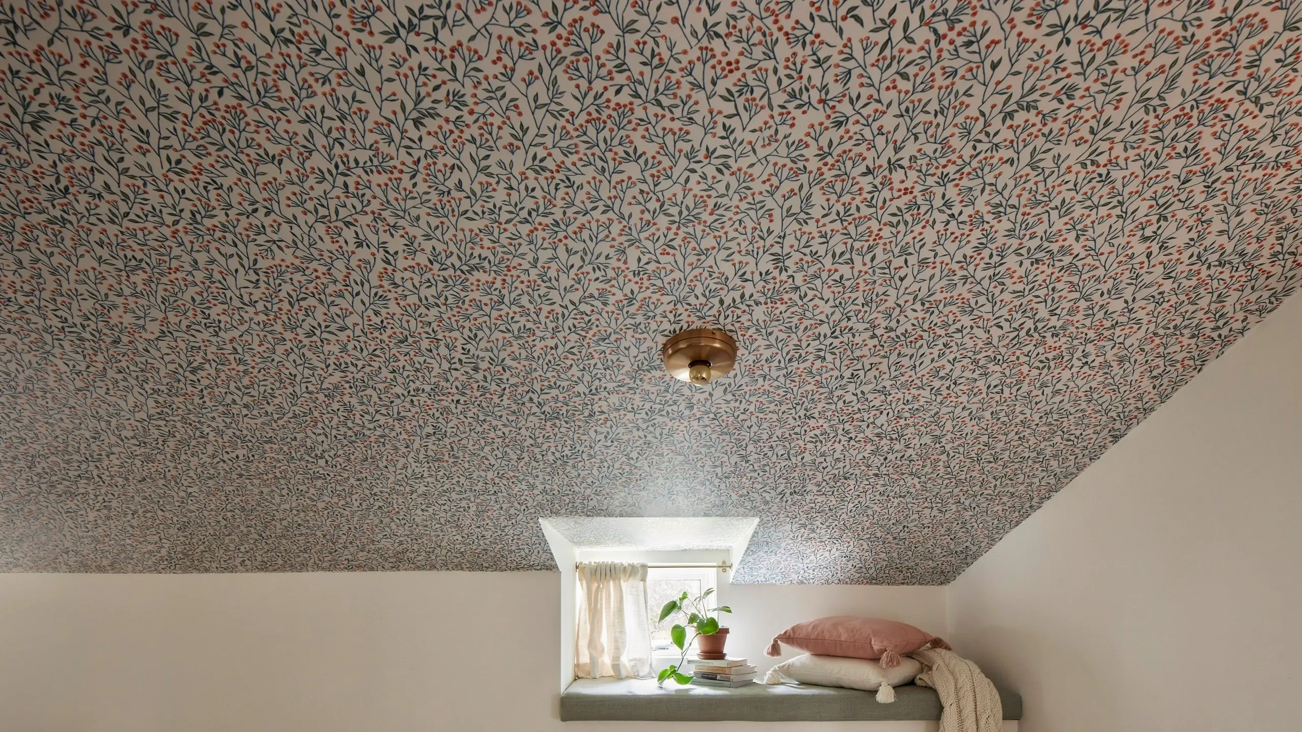 Attic bedroom featuring a sloped ceiling covered in white botanical wallpaper with green stems and small red berries, a brass flush-mount light, and a recessed window nook with sheer curtains, stacked books, a potted plant, and dusty pink and cream throw pillows resting on a grey window sill.
