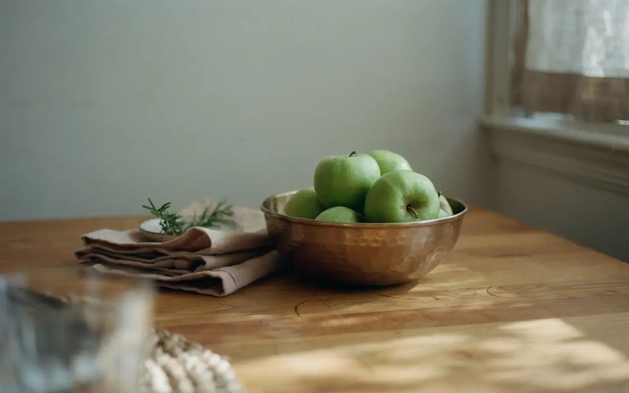 Brass bowl of green apples beside folded napkins on a warm wooden kitchen table.