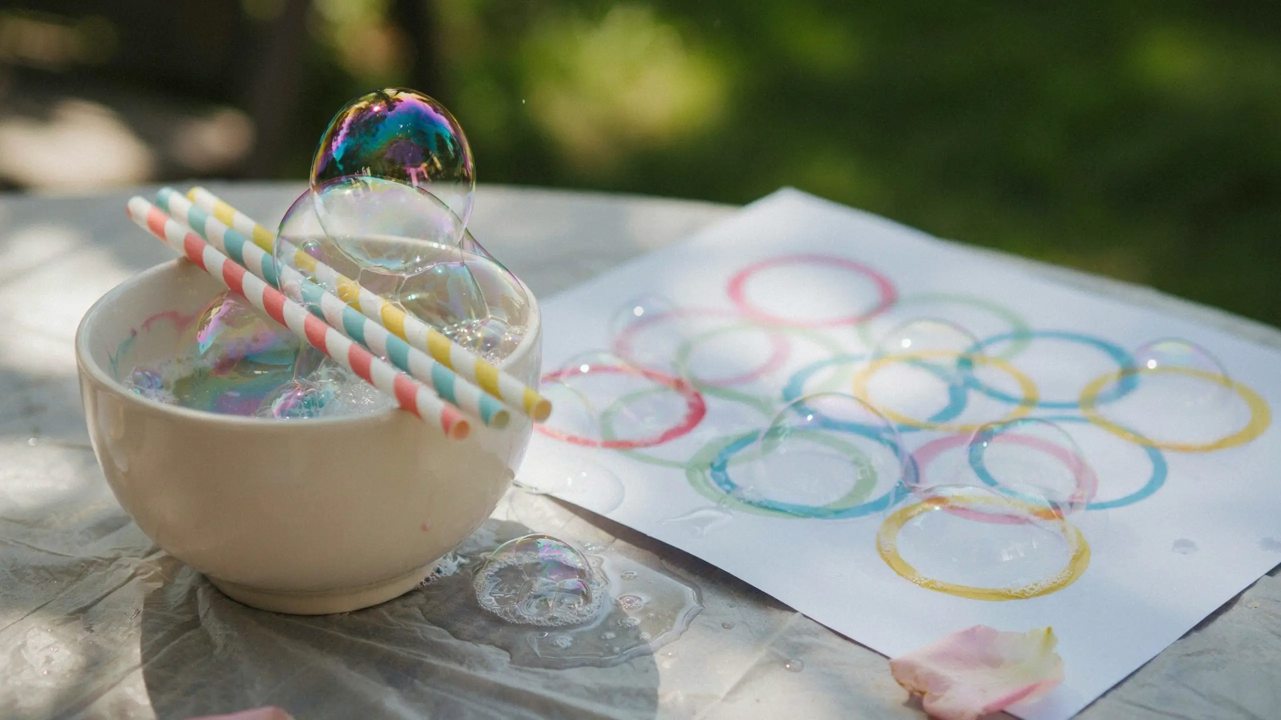 A close-up of a bubble print art craft setup outdoors, featuring a small white bowl of soapy solution with colorful striped straws and iridescent bubbles, sitting next to a white paper covered in overlapping rainbow-colored paint rings.