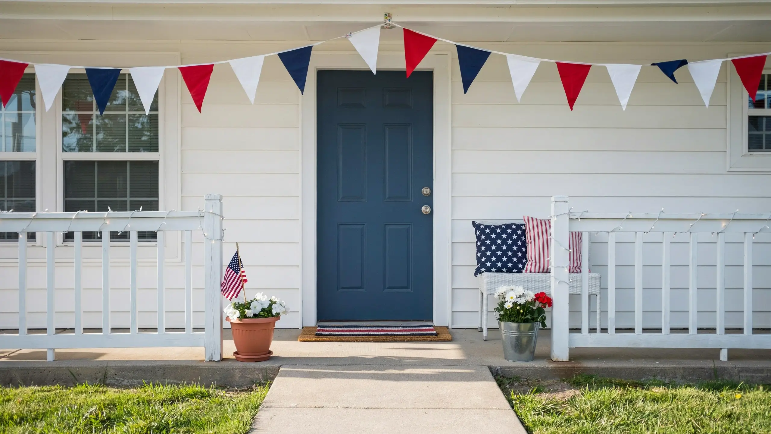 Budget-friendly July 4th porch with patriotic bunting, flag pillows, potted flowers and string lights.