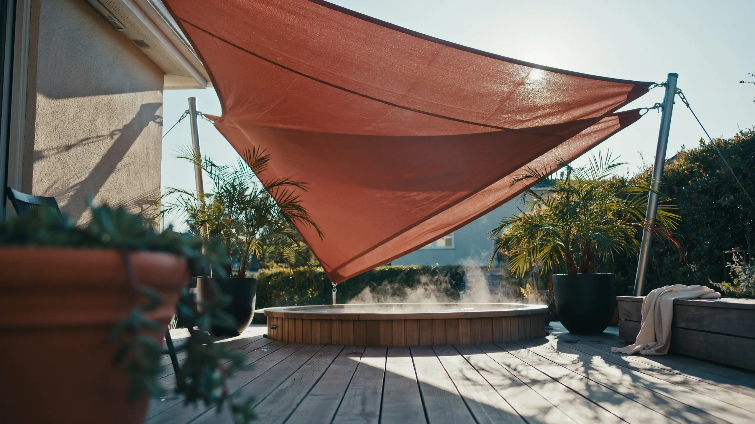 Large burnt orange shade sail providing overhead cover for a steaming wooden hot tub on a sunny backyard deck with potted tropical plants